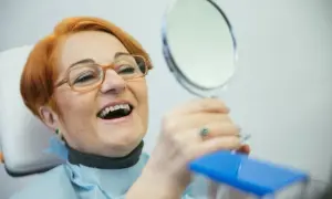 Uśmiechnięta kobieta z rudymi włosami ogląda swoje nowe zęby w lusterku. NFZ dofinansowanie do protez zębowych pozwoliło jej odzyskać pewność siebie.