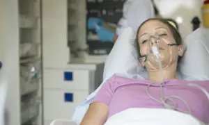 Młoda kobieta z maską tlenową na twarzy, leżąca w łóżku szpitalnym. W tle widać personel medyczny. Może to być objaw choroby popromiennej.