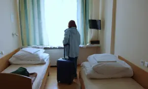 Kobieta z walizką przy oknie w pokoju sanatoryjnym. Zastanawia się, ile trzeba dopłacić do sanatorium.