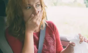 Kobieta w samochodzie zakrywa usta dłonią, trzymając chusteczkę. Może potrzebuje diphenhydramine na alergię.