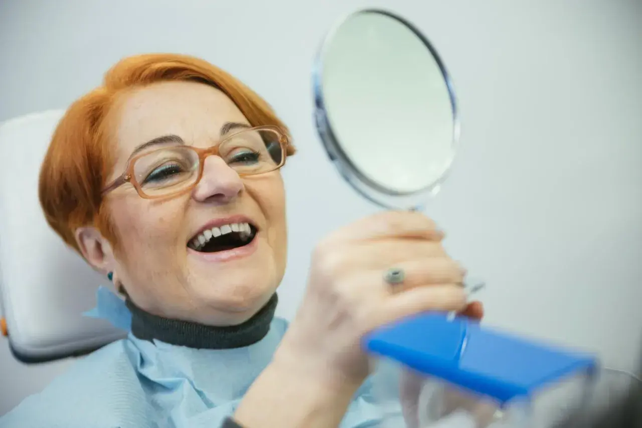 Uśmiechnięta kobieta z rudymi włosami ogląda swoje nowe zęby w lusterku. NFZ dofinansowanie do protez zębowych pozwoliło jej odzyskać pewność siebie.