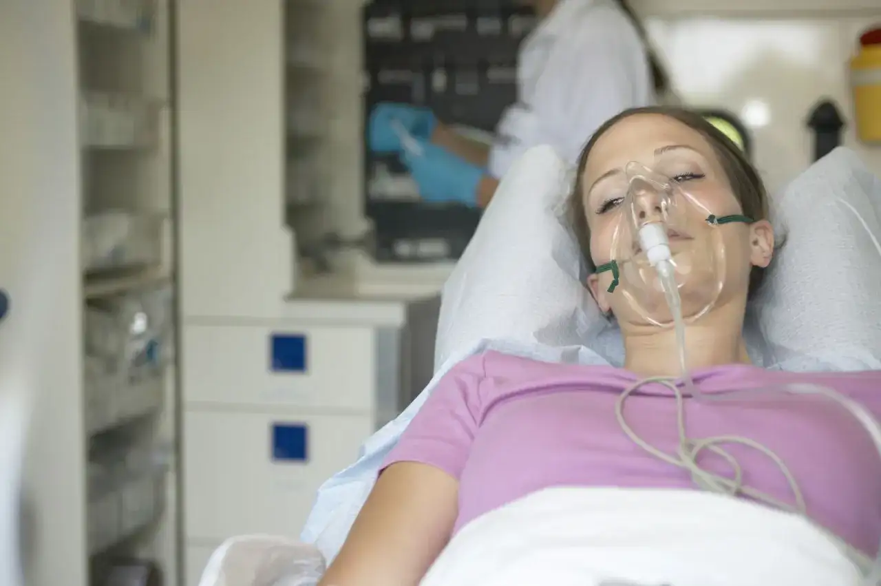Młoda kobieta z maską tlenową na twarzy, leżąca w łóżku szpitalnym. W tle widać personel medyczny. Może to być objaw choroby popromiennej.