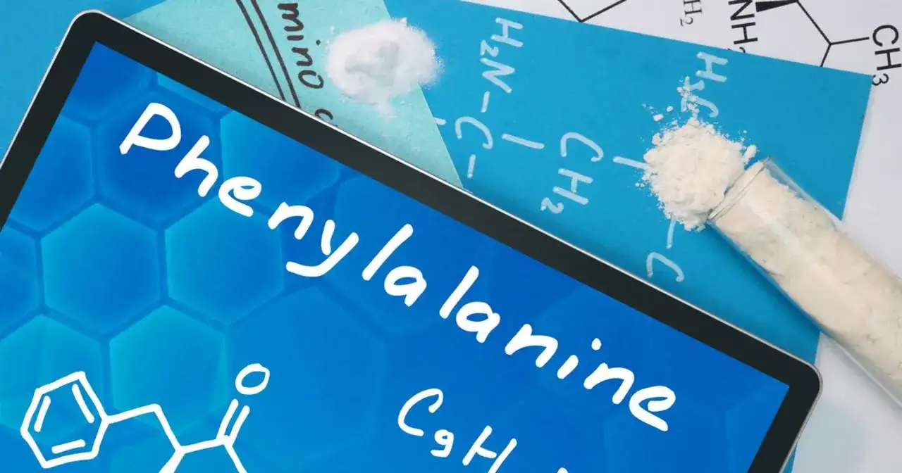 Tablet z napisem fenyloalanina i wzorem chemicznym. Obok probówka z białym proszkiem.