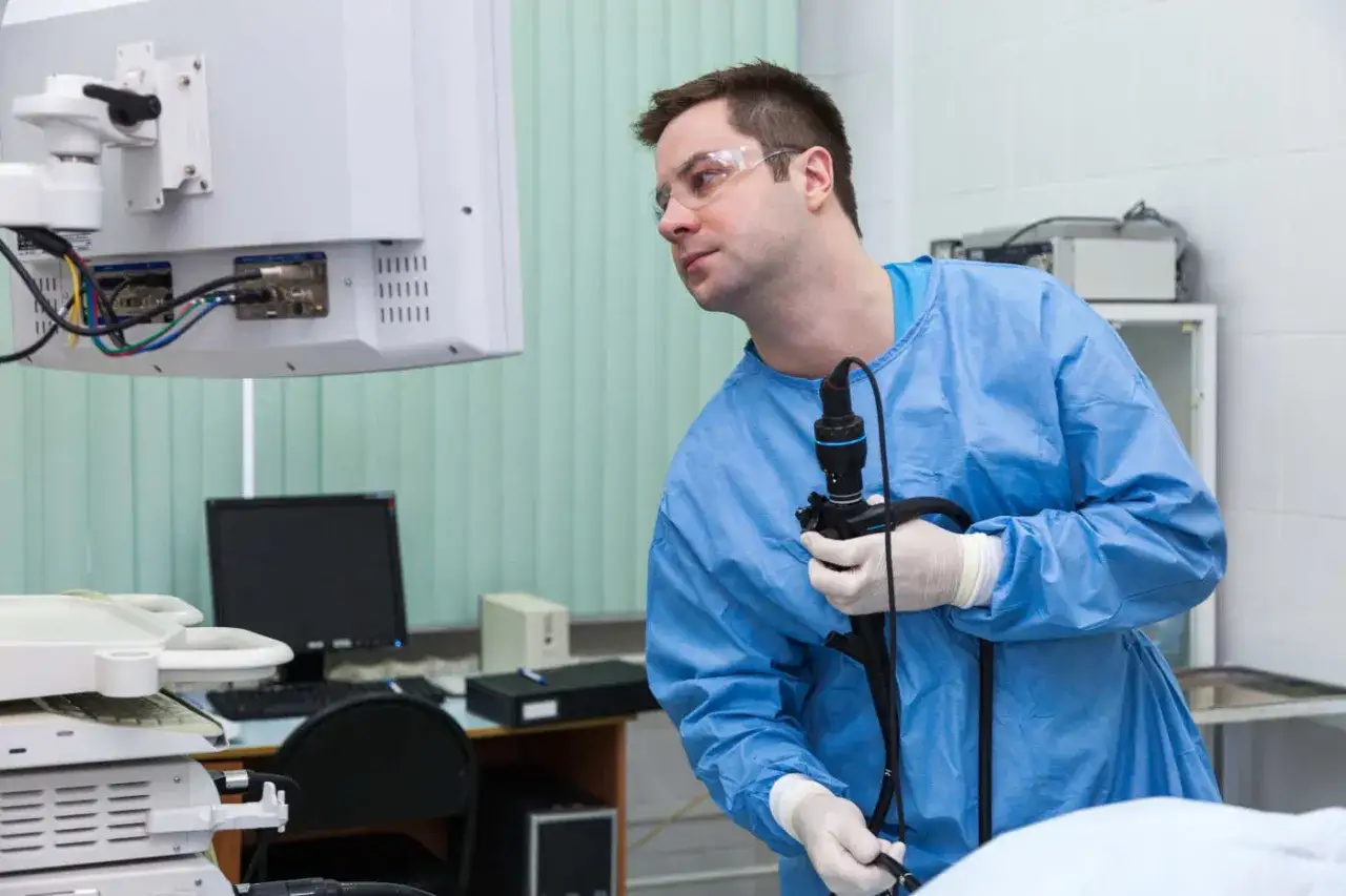 Lekarz w niebieskim fartuchu przygotowuje się do gastroskopii ze znieczuleniem NFZ, trzymając endoskop i patrząc na monitor.