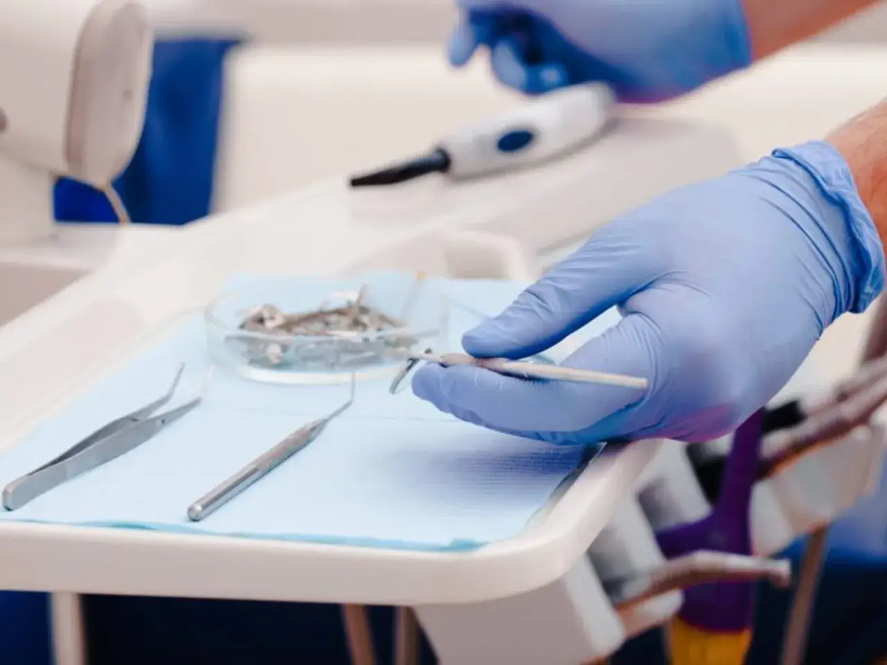 Dłoń w niebieskiej rękawiczce chwyta narzędzie dentystyczne. Zadbaj o swoje zęby, sprawdź gwarancję na protezy zębowe NFZ.