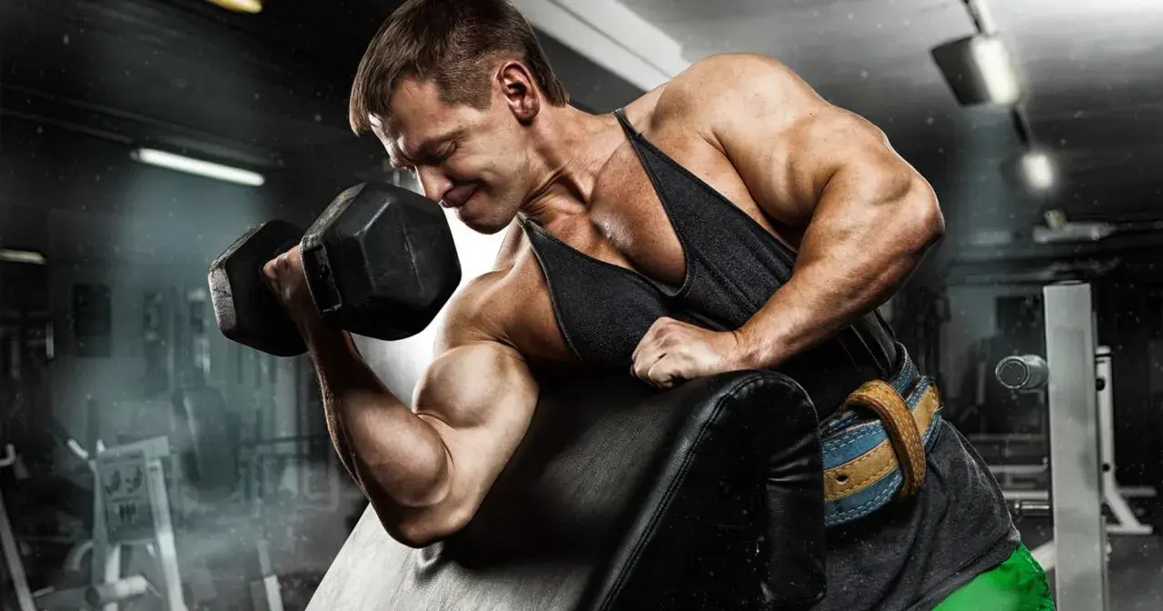 Mężczyzna ćwiczy biceps z hantlem, napinając mięśnie. W tle siłownia.