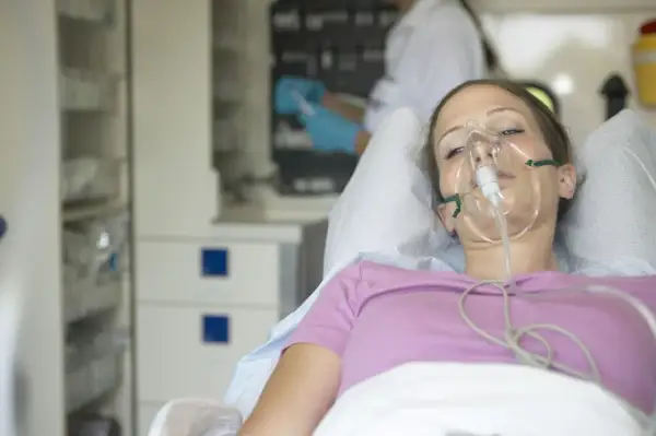 Młoda kobieta z maską tlenową na twarzy, leżąca w łóżku szpitalnym. W tle widać personel medyczny. Może to być objaw choroby popromiennej.