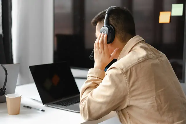 Mężczyzna w słuchawkach z mikrofonem, skupiony na pracy przy laptopie. Jego dłoń dotyka ucha, co może sugerować intensywne przetwarzanie słuchowe podczas rozmowy lub nauki.